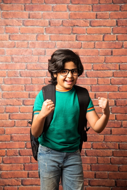 Retrato de niño asiático indio estudiante en ropa casual con mochila o mochila, de pie contra la pared de ladrillo rojo