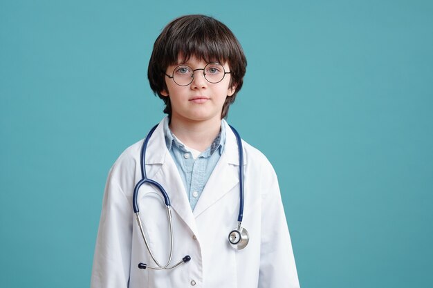 Retrato de niño en bata blanca jugando en doctor mirando a la cámara contra el fondo azul.