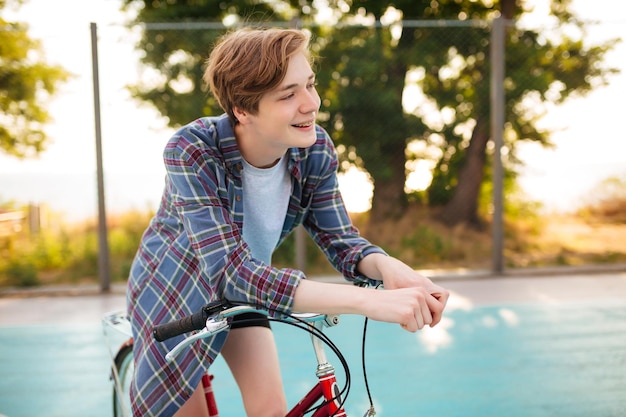 Retrato de niño con cabello rubio en camisa casual de pie con bicicleta roja en la cancha de baloncesto en el parque Joven mirando alegremente a un lado mientras está de pie con bicicleta clásica