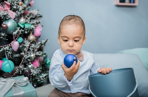 Retrato de un niño con una camisa azul con un globo de juguete sentado en una cama contra un árbol de Navidad