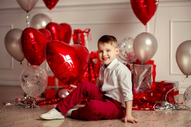 Retrato de un niño con una camisa blanca y pantalón rojo en el fondo de la decoración festiva de bolas rojas en el widget de corazones y regalos. cumpleaños de vacaciones o día de san valentín