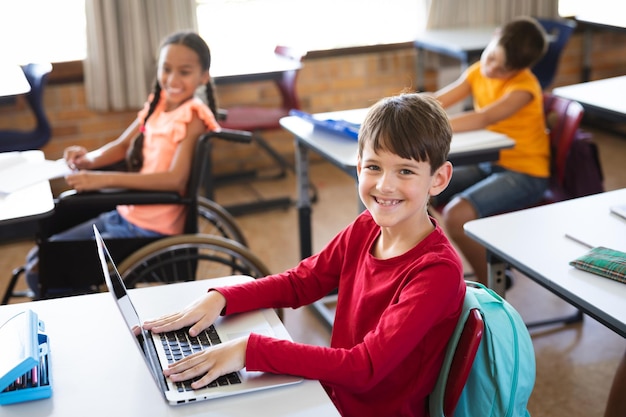 Retrato de un niño caucásico con una laptop sonriendo mientras se sienta en su escritorio en la escuela primaria