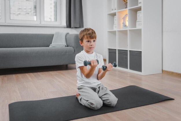 Retrato de niño caucásico niño haciendo ejercicio con pesas mientras está sentado en el piso de la sala de estar