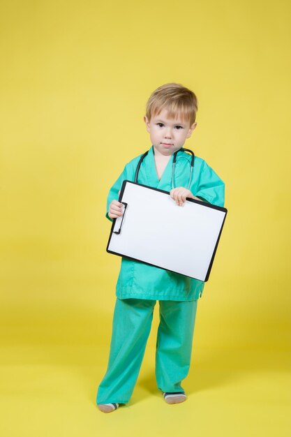 Retrato de un niño caucásico vestido con un abrigo verde de doctor sostiene una tableta en la mano aislada en amarillo
