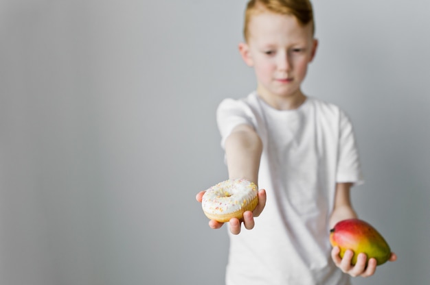 Retrato de un niño chico confundido eligiendo entre donut y mango sobre fondo gris