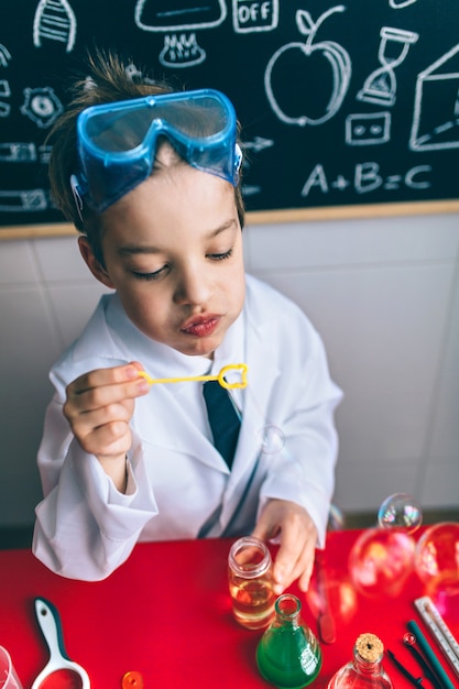 Retrato de niño científico jugando con pompas de jabón sobre la mesa contra la pizarra dibujada