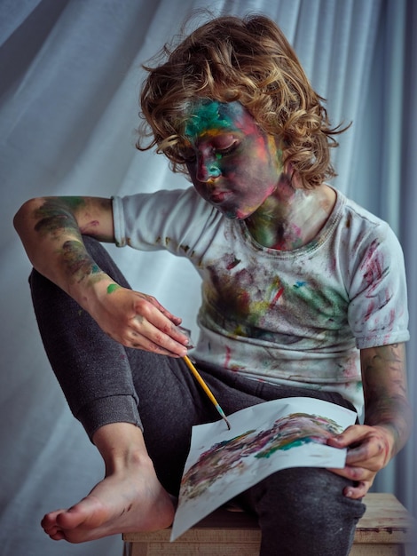 Retrato de un niño concentrado con la cara pintada, cuello y brazos, vistiendo una camiseta con manchas coloridas y pantalones mirando el papel colocado sobre las rodillas y pintando un cuadro