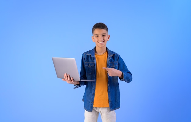 Retrato de un niño confiado sonriendo y señalando a la computadora portátil mientras trabaja sobre un fondo azul aislado