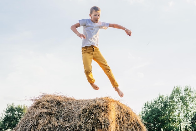 Retrato de un niño descalzo con pantalones cortos blancos y amarillos saltando sobre un pajar en el campo. Actividad al aire libre. Vista lateral