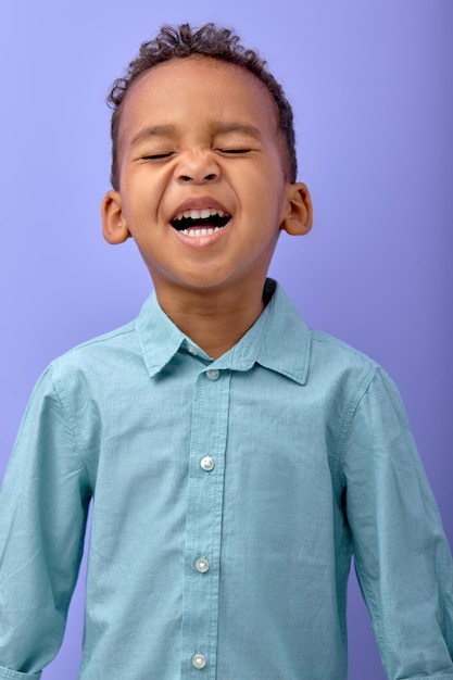 Foto retrato de niño emocionalmente negro. niño divertido aislado sobre fondo púrpura. niño rizado en camisa diviértete, emocionado, riendo, posando en cámara, foto de estudio. emociones humanas, concepto de niños