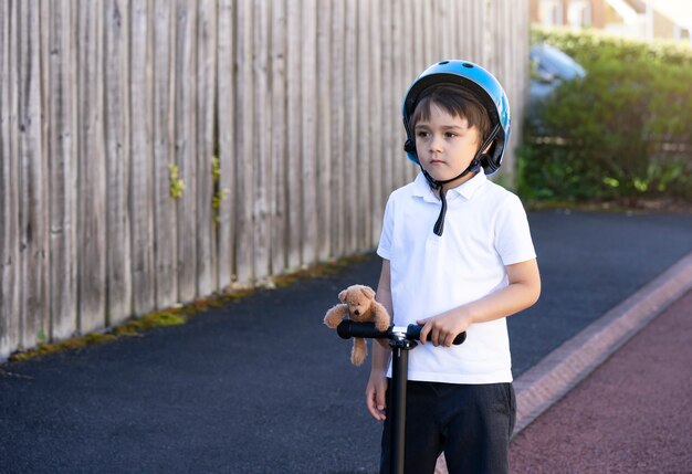 Retrato niño de escuela montar un scooter a la escuela, Niño con casco de seguridad montando un rodillo, Niño de pie con oso de peluche mirando en lo profundo, Ocio activo y deporte al aire libre para niños.