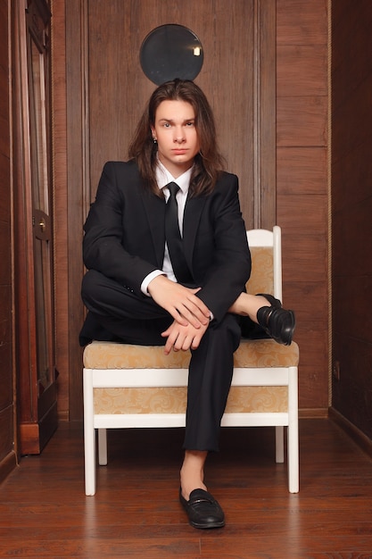 Foto retrato de niño de escuela. vestido formalmente con traje negro, corbata, camisa blanca, las manos en los bolsillos, un joven y guapo estudiante universitario de pie contra la puerta de la oficina de estilo vintage en el campus, mirándote.