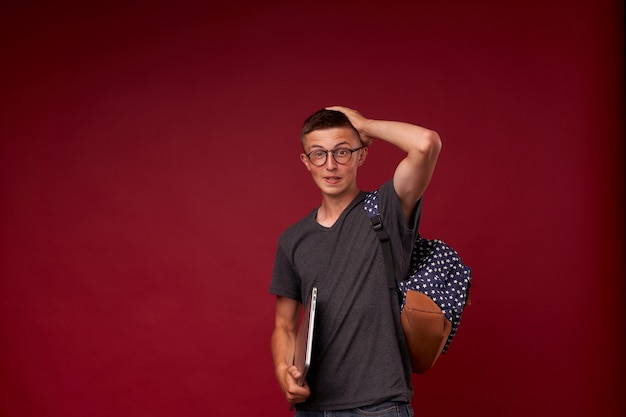 Retrato de un niño estudiante con una mochila y una computadora portátil en sus manos sonriendo en rojo