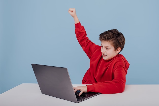 Retrato de niño exitoso ganador con manos levantadas niño en sudadera roja cerca de la mesa con computadora portátil ...