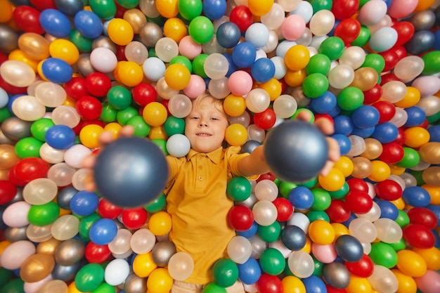 Retrato de niño feliz acostado entre bolas de colores y lanzando las bolas