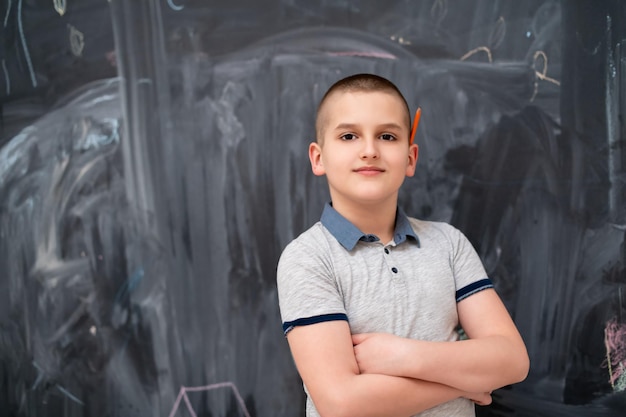 retrato de un niño feliz con un bolígrafo de madera naranja detrás de la oreja parado frente a una pizarra negra