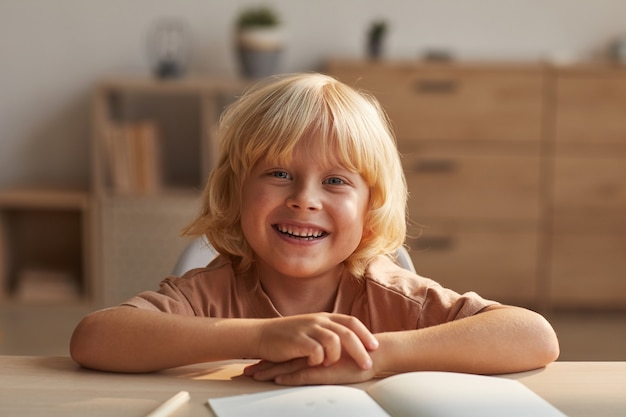 Retrato de niño feliz con cabello rubio sonriendo mientras está sentado en la mesa con cuadernos