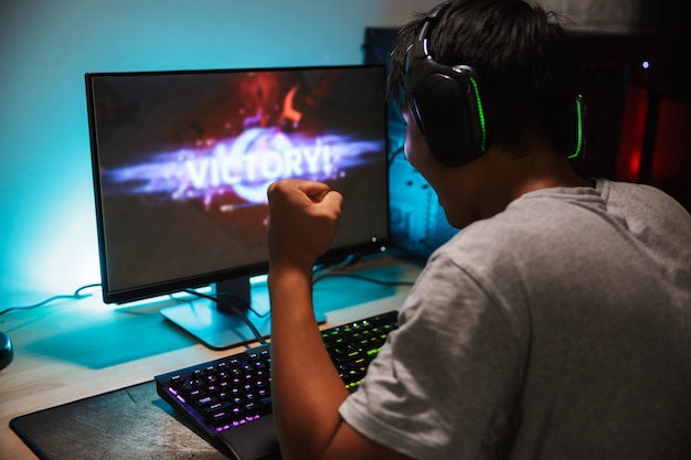 Retrato de niño feliz jugador adolescente ganando mientras juega videojuegos en la computadora en una habitación oscura, usando audífonos y usando un teclado colorido retroiluminado