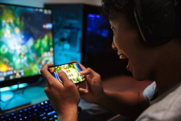 Retrato de niño feliz jugador adolescente jugando videojuegos en el teléfono inteligente y la computadora en una habitación oscura, usando audífonos y usando un teclado colorido retroiluminado