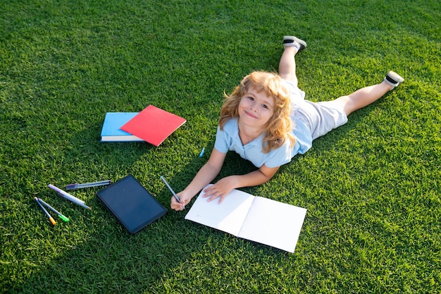 Retrato de un niño feliz con un libro escrito en un cuaderno en el parque Educación temprana para niños Un niño pequeño lee un libro en el jardín Tarea de vacaciones de verano Estudiante de preescolar al aire libre