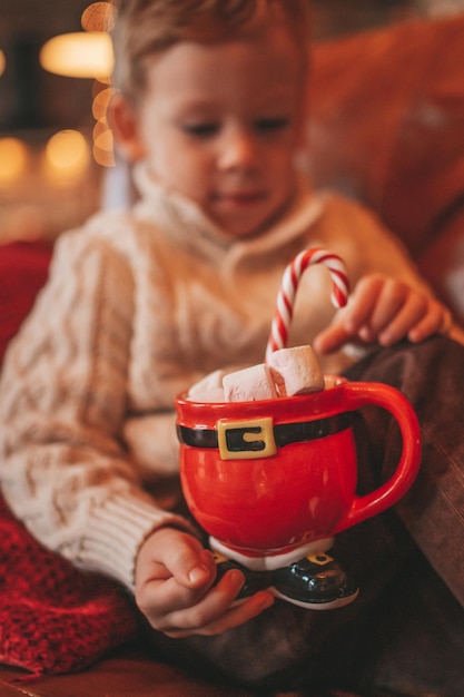 Retrato de un niño feliz sincero en un suéter beige de punto que sostiene una taza de Navidad con malvaviscos y bastón de caramelo