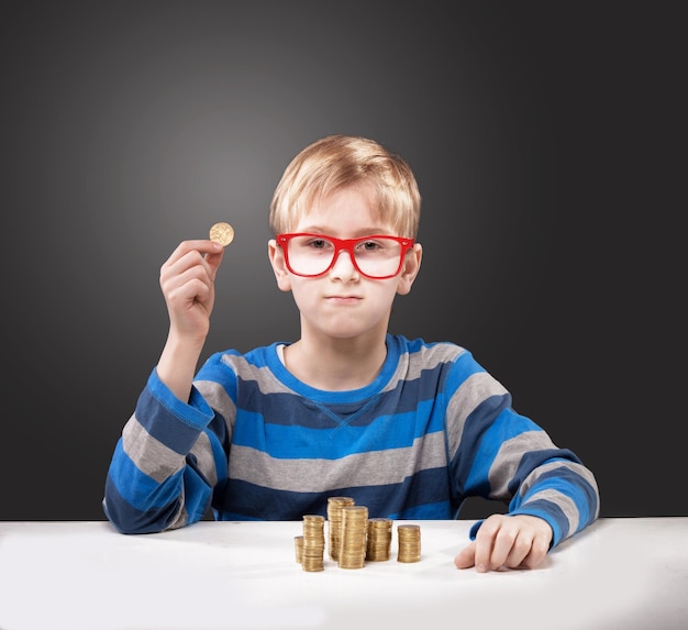 Retrato de un niño con gafas enmarcadas en rojo con una moneda en la mano mirando a la cámara