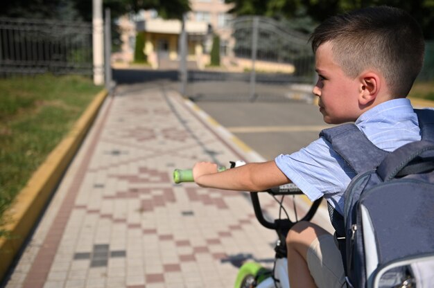 Foto retrato de niño guapo con mochila escolar montando bicicleta al establecimiento escolar. niño regresando a la escuela. vista desde atrás