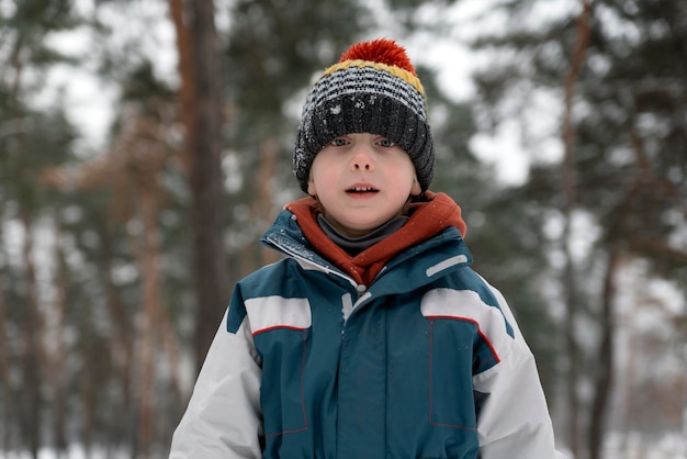 Retrato de niño lindo en el bosque de invierno sobre fondo de árboles Niño con gorro de punto y chaqueta de invierno mira directamente a la cámara