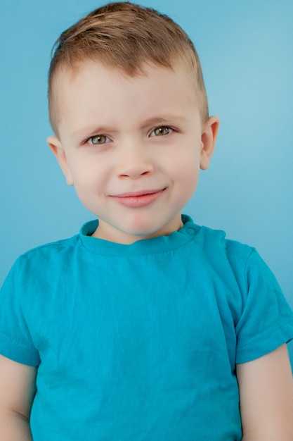 Retrato de niño lindo con elegante peinado rizado en camiseta azul de pie, mirando a la cámara con cara seria y atenta, tranquila expresión pensativa.