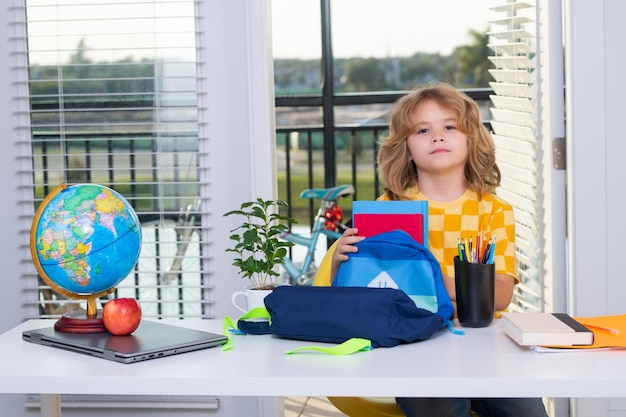 Retrato de niño lindo niño escolar sentado en la mesa en casa pone útiles escolares en una mochila pr