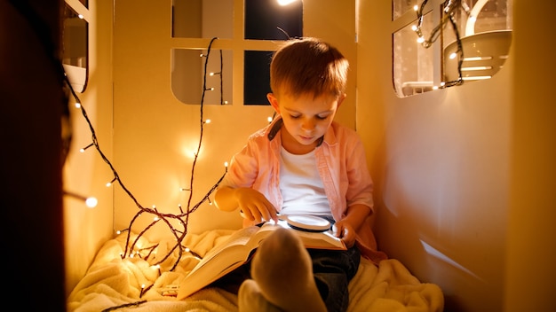 Retrato de niño con linterna jugando en su casa de juguete por la noche y leyendo un libro grande. Concepto de educación infantil y lectura en una habitación oscura.