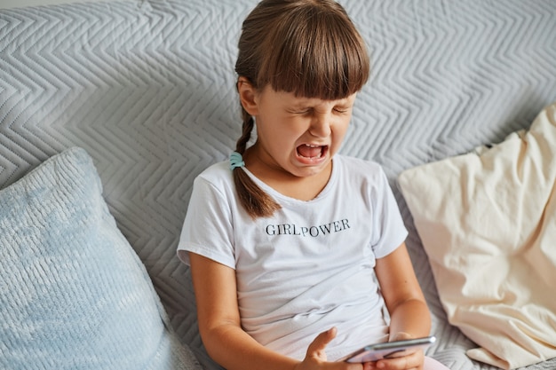 Retrato de niño llorando lindo molesto con cabello oscuro y coletas, sentado en el sofá con el teléfono celular en las manos, triste por perder nivel en su juego favorito, con camiseta blanca.