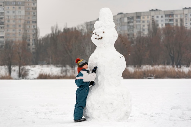 Retrato de niño y muñeco de nieve enorme. Niño esculpe muñeco de nieve en invierno cubierto de nieve a pie con el telón de fondo de edificios de gran altura.