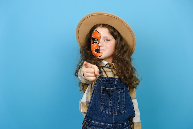 Retrato de niño niña con máscara de maquillaje de Halloween lleva sombrero y camisa, señalar con la cámara del dedo índice en su motivación, posando sobre fondo de color azul en estudio. Concepto de fiesta de vacaciones