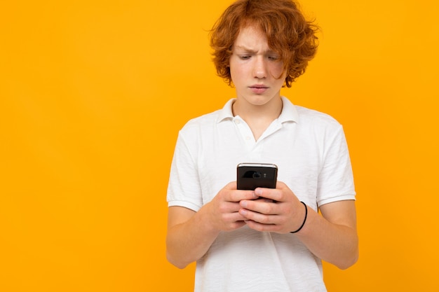 Retrato de un niño pelirrojo en una camiseta blanca con un teléfono en sus manos en amarillo con espacio de copia