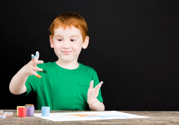 Retrato de un niño pelirrojo feliz dibuja en papel con pinturas de diferentes colores, el niño dibuja con sus manos y por eso sus manos están pintadas en azul, rojo y otros colores