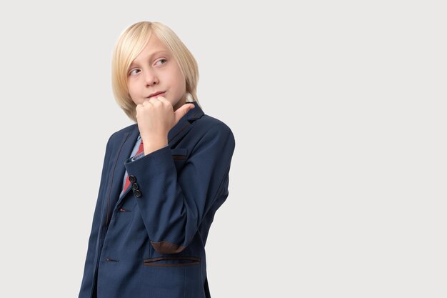 Retrato de un niño pensativo con cabello rubio mantiene la mano en la barbilla reflexiona sobre algo vestido con un traje azul oscuro aislado sobre un fondo blanco Gente y concepto de negocios