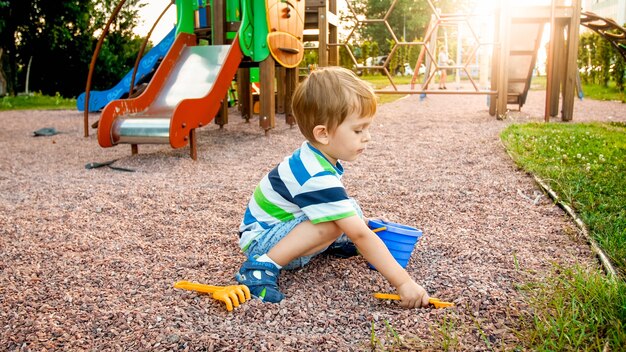 Retrato de niño pequeño de 3 años sentado en el patio de recreo y cavando arena con pala de plástico de juguete