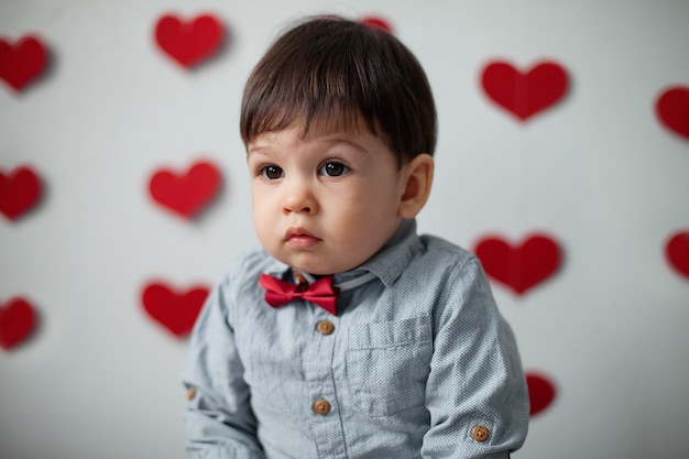 Retrato de un niño pequeño en una camisa con una pajarita sobre un fondo de pared con corazones en el día de San Valentín.
