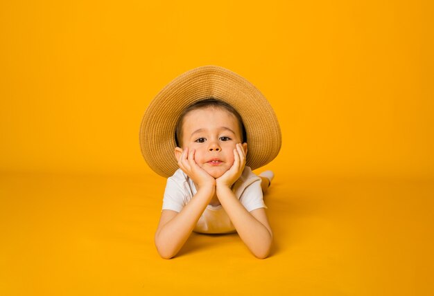 Retrato de un niño pequeño con una camiseta blanca y un sombrero de paja mirando a la cámara sobre una superficie amarilla con espacio para texto