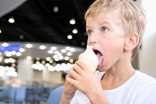 Retrato de un niño pequeño y divertido hambriento comiendo delicioso helado frío en una taza de gofres sentado en un café de fondo oscuro