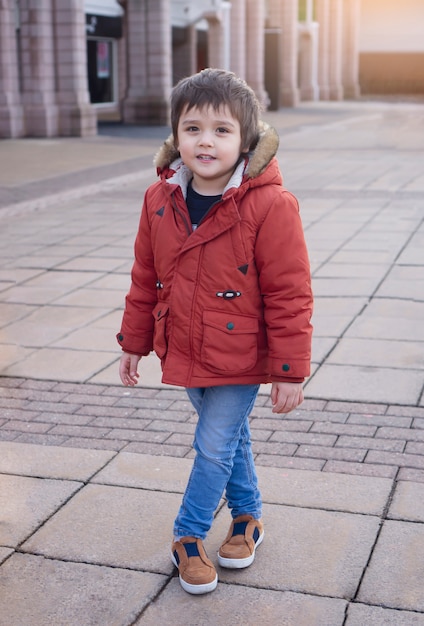 Retrato del niño pequeño feliz que se coloca en parque de la ciudad en tono retro, niño activo que mira la cámara con la cara sonriente mientras que se coloca fuera de centro comercial.