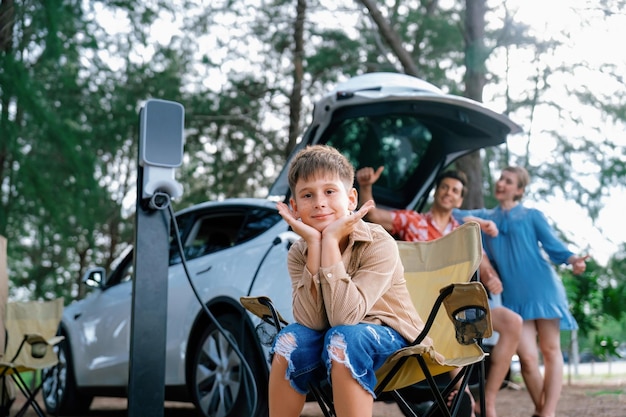 Retrato de niño pequeño sentado en una silla de camping con su familia en el fondo Viaje por carretera con estación de carga de energía alternativa para el concepto de automóvil ecológico Perpetuo