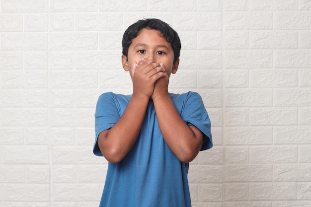 Retrato de un niño pequeño sorprendido que cubre la boca con las manos sobre fondo blanco.