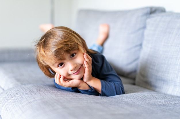 Retrato de niño preescolar acostado en un sofá gris claro, mirando a la cámara con una sonrisa. Niño que pasa tiempo en casa.