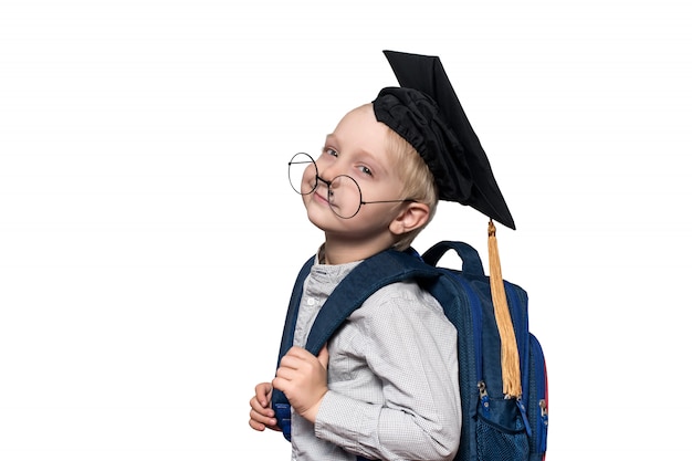 Retrato de un niño rubio cansado en gafas, un sombrero académico y una mochila. Concepto de escuela Aislar