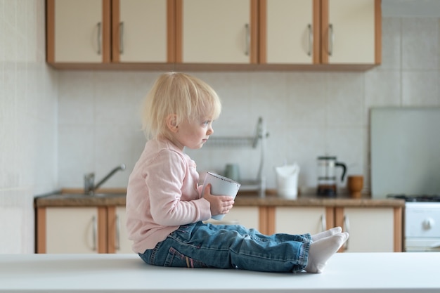 Retrato de niño rubio en cocina con taza en sus manos. Vista lateral.