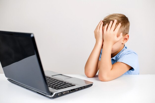 Retrato de niño rubio en la computadora portátil que cubre su rostro con las manos con miedo