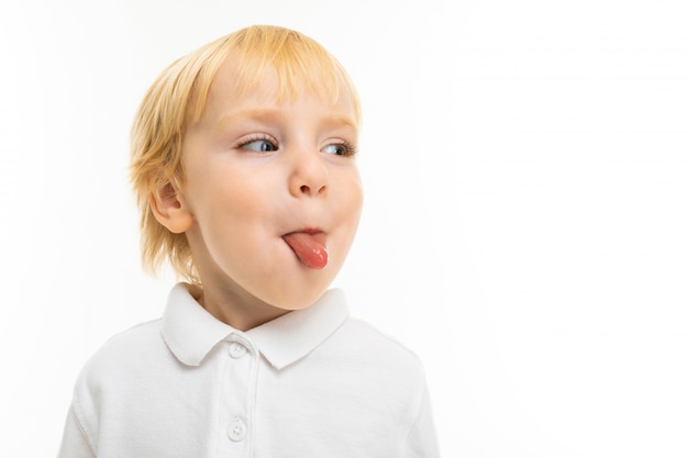 Retrato de niño rubio europeo vistiendo una camiseta blanca que muestra la lengua en una pared blanca del estudio con espacio en blanco