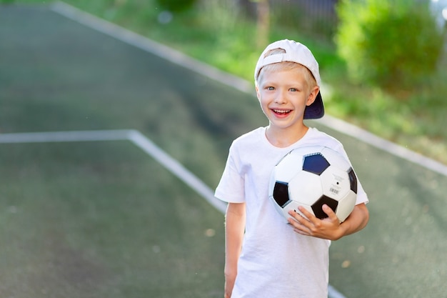 Retrato de un niño rubio con una gorra en un uniforme deportivo con un balón de fútbol en sus manos en el campo de fútbol